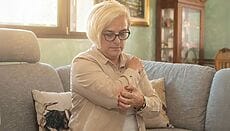 Ritratto fotografico realistico verticale, luce calda naturale del pomeriggio, persona adulta davanti a una finestra con tenda chiara, mano che sfiora l’avambraccio vicino al gomito come per calmare il prurito, mood intimo e rassicurante, pelle solo con lieve arrossamento, sfondo casa sfocato, colori naturali, profondità di campo leggera, niente set da studio, nessun testo, nessun logo, niente dettagli grafici